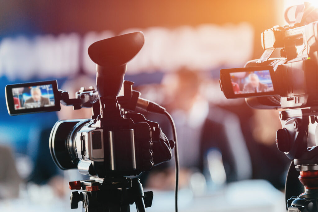Two,Cameras,Recording,Presentation,At,Press,Conference,,Blurred,Speakers,Wearing webcast equipment